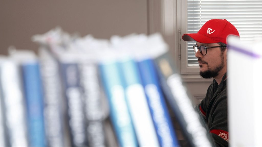 Berthold in the background writing while a book shelf is in the forefront